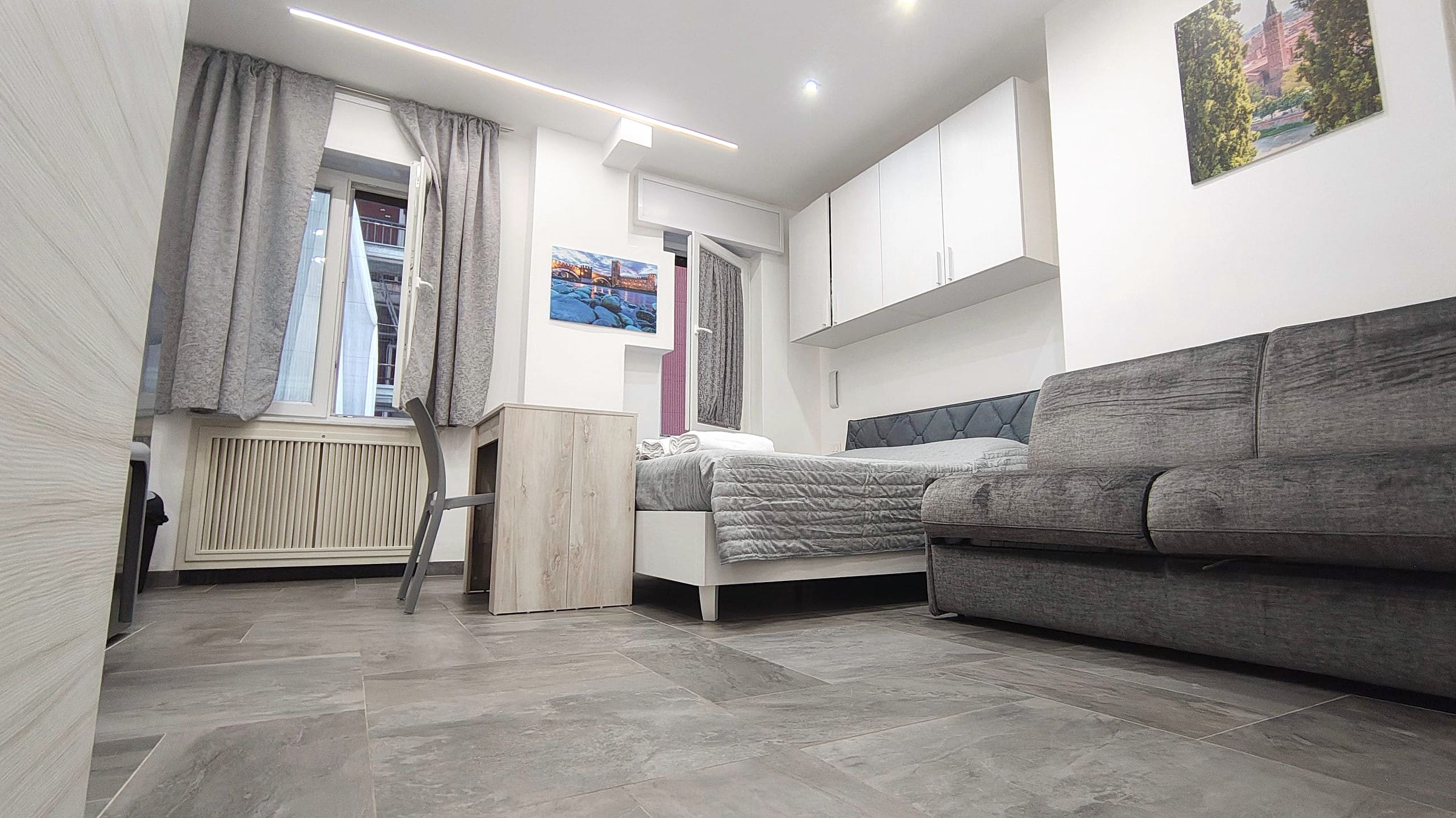 Camera quadrupla vista dal basso: letto, divano, scrivania, finestra con tenda e pensili bianchi